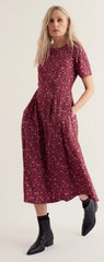 A woman modelling Seasalt short sleeved midi dress with a dense small floral print in burgundy and pink, paired with black ankle boots, against a plain light background.