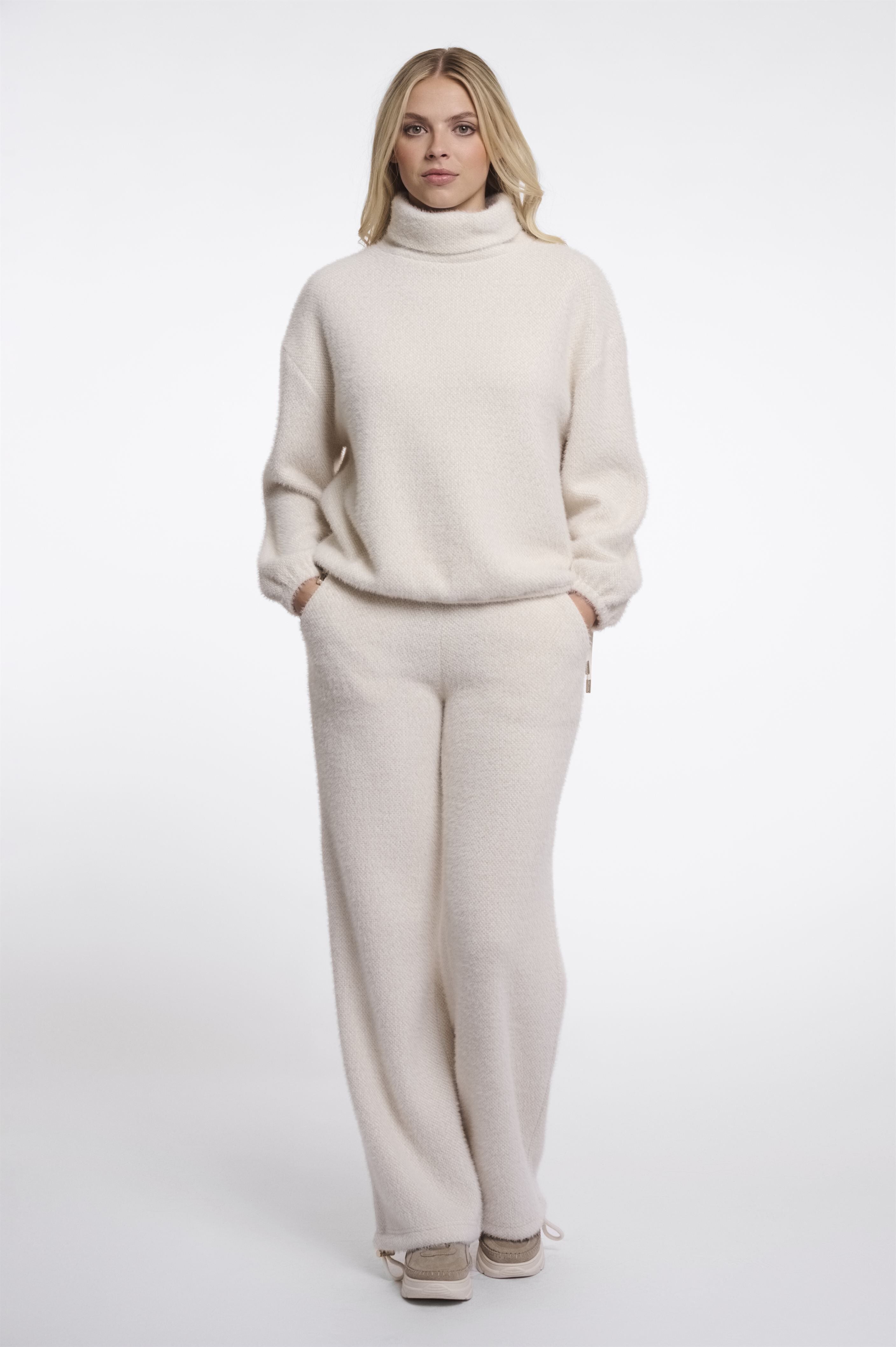 A woman modeling Rino & Pelle cream turtleneck jumper with matching wide-leg trousers, styled with beige sneakers against a plain white studio background.