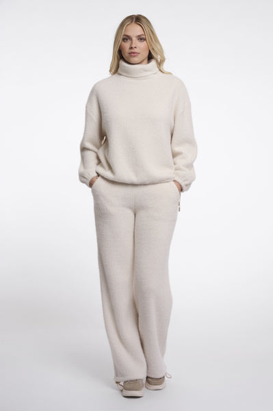 A woman modeling Rino & Pelle cream turtleneck jumper with matching wide-leg trousers, styled with beige sneakers against a plain white studio background.