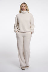 A woman modeling Rino & Pelle cream turtleneck jumper with matching wide-leg trousers, styled with beige sneakers against a plain white studio background.