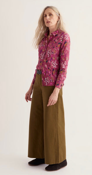 A woman modelling Seasalt a long-sleeve button-up shirt with a pink and purple floral pattern, paired with wide-leg olive brown trousers and dark slip-on shoes, against a plain off-white studio background.
