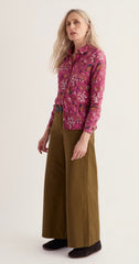 A woman modelling Seasalt a long-sleeve button-up shirt with a pink and purple floral pattern, paired with wide-leg olive brown trousers and dark slip-on shoes, against a plain off-white studio background.