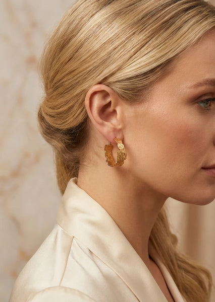A close-up side profile of a blonde woman wearing Envy textured gold hoop earring with a small gold disc charm, paired with a cream satin jacket against a soft beige marble background.