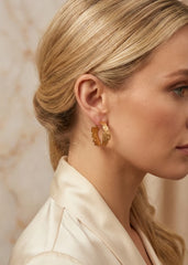 A close-up side profile of a blonde woman wearing Envy textured gold hoop earring with a small gold disc charm, paired with a cream satin jacket against a soft beige marble background.