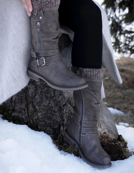 A person wearing Lunar Footwear taupe brown knee high boots with buckle straps and side zippers, seated outdoors in snow beside a tree trunk.
