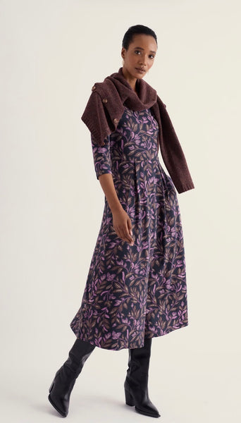A model wearing a Seasalt dark purple and black floral print midi dress with three-quarter sleeves, styled with a brown faux-fur collar and a matching brown jacket draped over her shoulders, paired with black pointed-toe boots against a light beige studio background.