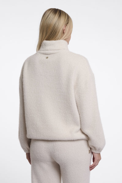 A woman viewed from the back wearing a fuzzy cream-coloured roll neck sweater and matching sweatpants against a plain white studio background.