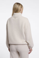 A woman viewed from the back wearing a fuzzy cream-coloured roll neck sweater and matching sweatpants against a plain white studio background.