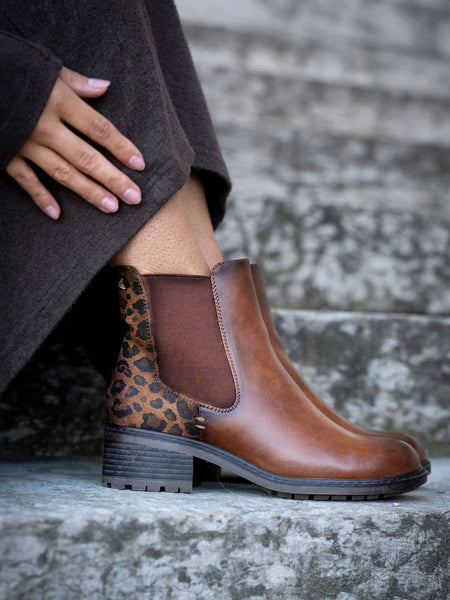 A person seated on stone steps wearing a brown Lunar Women's Chelsea boot with a leopard-print heel panel and dark elastic side gore.