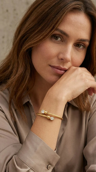 A close-up shot of a woman with brown hair resting her chin on her hand, wearing an Envy gold cuff bracelet with a small clear gemstone on her wrist. She is wearing a beige satin button-up shirt against a neutral textured background.