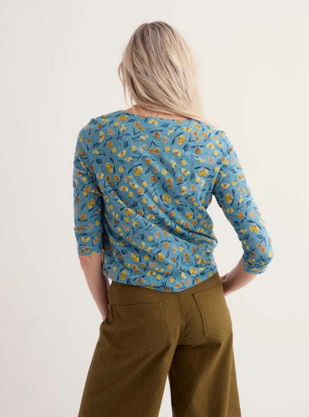 A woman shown from the back wearing a Seasalt  blue three quarter sleeve top with a yellow and green floral pattern, paired with olive green pants, against a plain off-white background.
