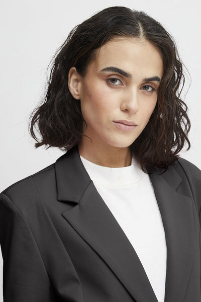 A woman wearing a black blazer over a white crewneck top, photographed against a light grey studio background.