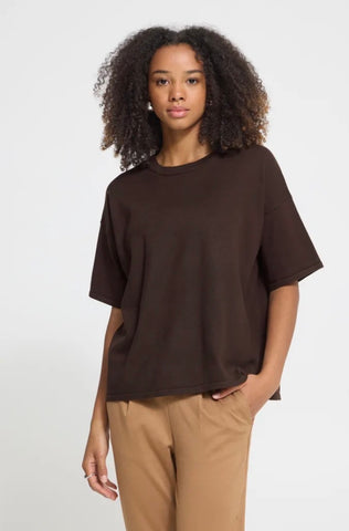 Close up of a person wearing an oversized dark brown top with short sleeves and a crew neckline, against a light grey background.