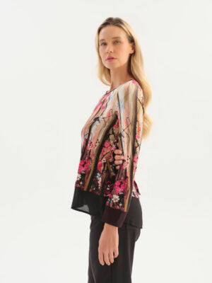 A woman modeling a long-sleeve Dolcezza blouse in vibrant pink floral paired with black trousers, against a plain white studio background.