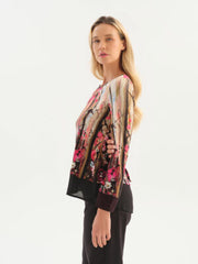 A woman modeling a long-sleeve Dolcezza blouse in vibrant pink floral paired with black trousers, against a plain white studio background.