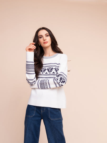A woman modelling Alison Sheri white knit sweater with white and navy geometric pattern across the chest and sleeves, paired with dark blue jeans, against a solid beige background.