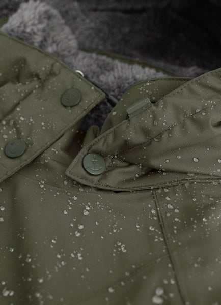 Close-up of an olive green waterproof jacket with water droplets on the surface, showing a buttoned flap and a gray faux-fur collar lining.