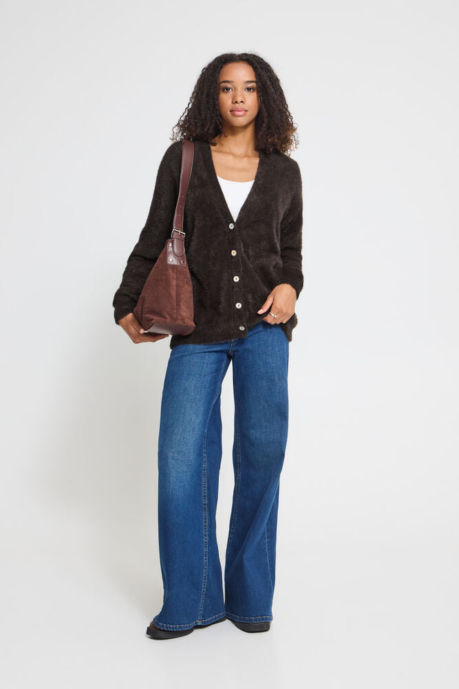 A woman stands against a light grey studio background wearing a Ichi dark brown  button-front cardigan over a white top, blue wide-leg jeans, and black shoes, while carrying a brown shoulder bag.