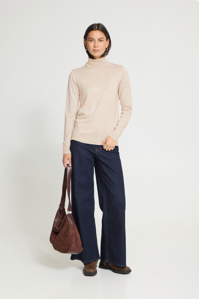 A woman stands against a light grey studio background wearing a light beige roll neck sweater, dark navy wide-leg trousers, and brown shoes, while holding a brown leather shoulder bag.