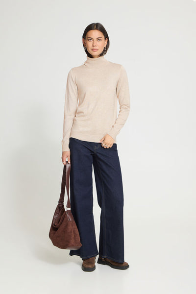 A woman stands against a light grey studio background wearing a light beige roll neck sweater, dark navy wide-leg trousers, and brown shoes, while holding a brown leather shoulder bag.