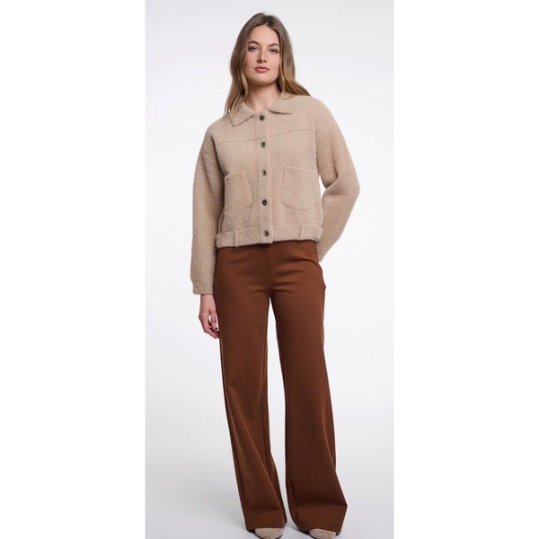 A woman modelling a Rino & Pelle cropped beige button-front jacket with front patch pockets, paired with brown wide-leg trousers, against a plain light grey studio background.