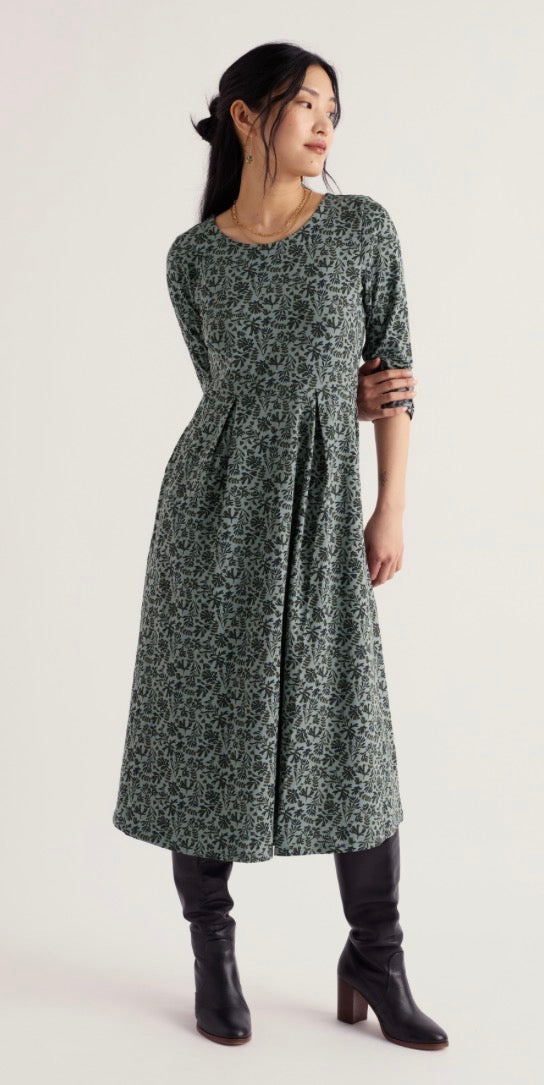 A woman modelling a Seasalt dark green and navy floral print midi dress with elbow-length sleeves, paired with black knee-high boots, against a plain off-white studio background.