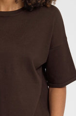 Close up of a person wearing an oversized dark brown top with short sleeves and a crew neckline, against a light grey background.
