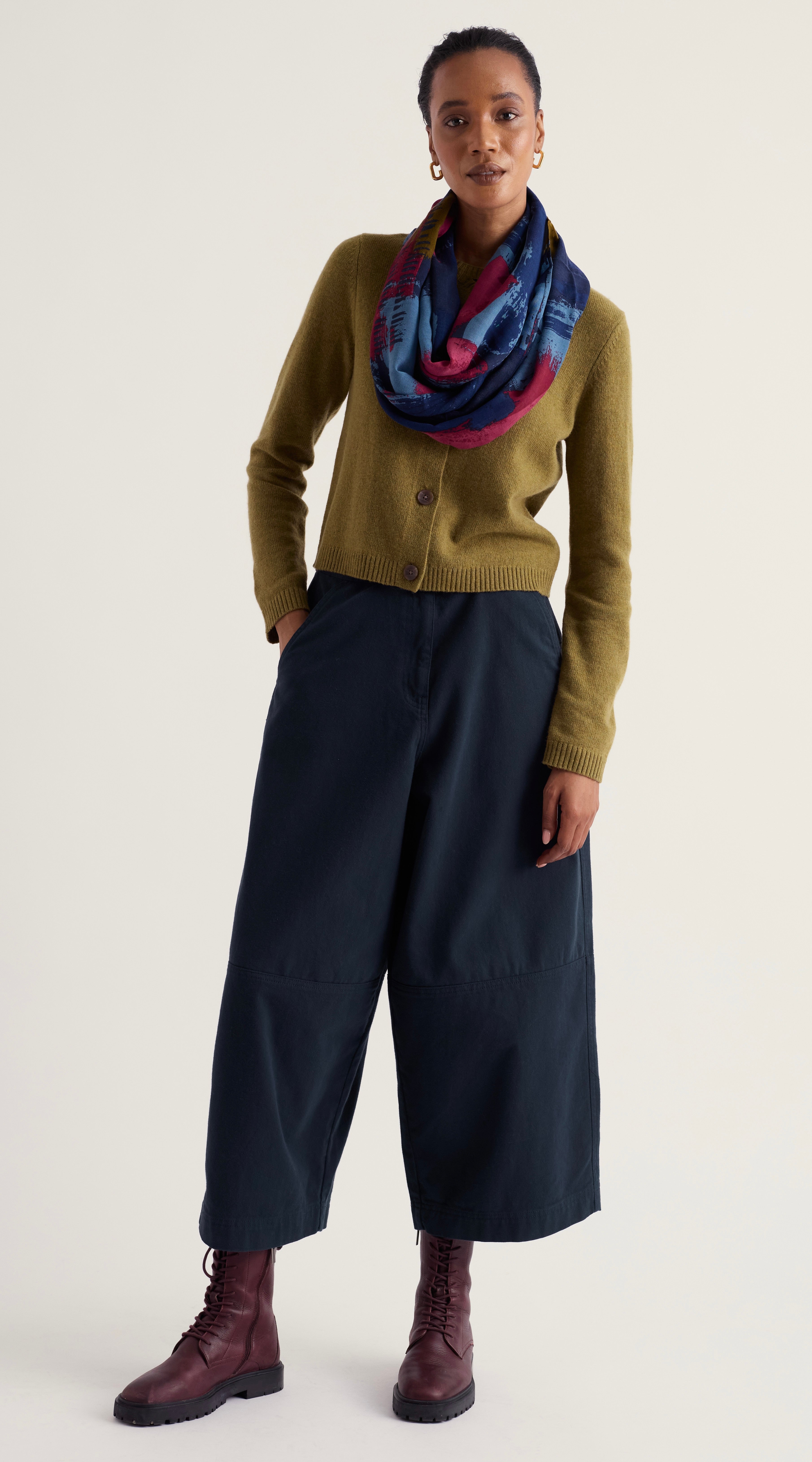 A model stands against a plain light background wearing Seasalt olive green cardigan, dark navy wide-leg cropped trousers, a blue and purple patterned infinity scarf, and dark brown leather lace-up boots.