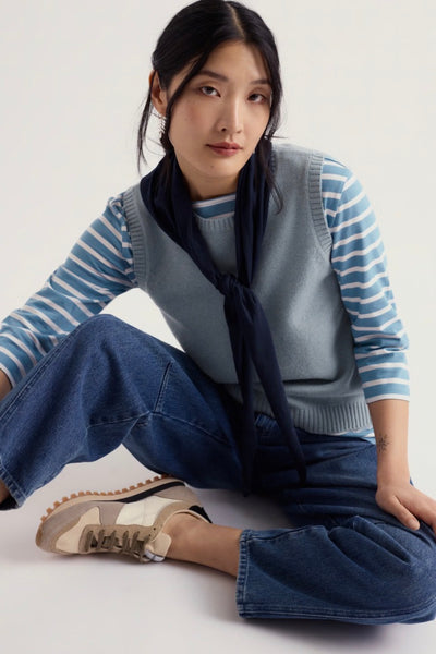 A woman sits cross legged on a light studio floor wearing Seasalt light blue vest top and a white striped long-sleeve top, a dark navy scarf tied at the neck, wide-leg blue jeans, and beige-and-white sneakers.