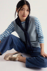 A woman sits cross legged on a light studio floor wearing Seasalt light blue vest top and a white striped long-sleeve top, a dark navy scarf tied at the neck, wide-leg blue jeans, and beige-and-white sneakers.