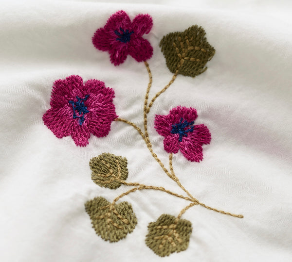 Close up of embroidered flowers and leaves on white fabric. The design features three magenta pink blossoms with dark blue centers and olive-green leaves connected by tan stems.