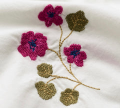 Close up of embroidered flowers and leaves on white fabric. The design features three magenta pink blossoms with dark blue centers and olive-green leaves connected by tan stems.