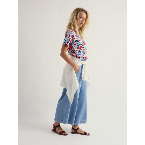 Woman wearing a floral top and blue pants with a white bag, standing against a plain background.