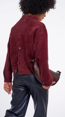 A model from the back wearing a Rino & Pelle, cropped maroon button-up jacket and shiny black leather pants, holding a brown leather shoulder bag against a plain white background.