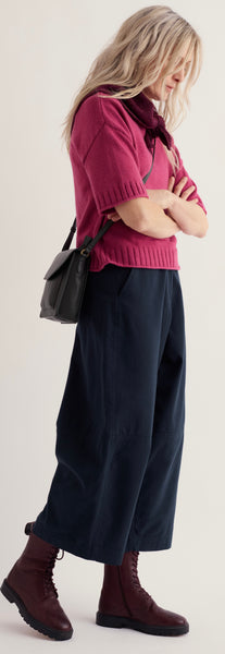 A woman modelling Seasalt short sleeve knit sweater in dusty rose pink, paired with dark navy wide leg trousers and dark brown lace up boots, carrying a small black shoulder bag against a light grey studio background.