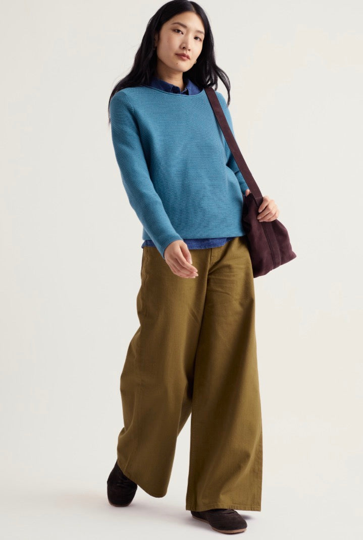A woman modelling Seasalt blue knit sweater, olive green wide-leg trousers, and a dark brown shoulder bag against a plain light background.