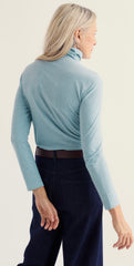 Back profile of woman wearing light blue long sleeve, roll neck top, with blue jeans against a white studio background.