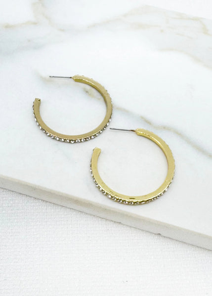 A pair of Envy gold hoop earrings with textured edges and small clear rhinestones, displayed on a white marble surface.
