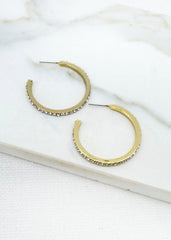 A pair of Envy gold hoop earrings with textured edges and small clear rhinestones, displayed on a white marble surface.