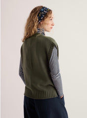 A woman shown from the back wearing an olive green knit vest over a navy and white striped turtleneck, paired with dark navy pants and a blue patterned headband, against a plain off-white background.