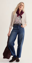 A woman modelling alight beige knit cardigan over a Seasalt floral top, paired with a dark burgundy scarf, wide-leg blue jeans, dark socks, and brown shoes, holding a dark brown tote bag against a plain off white background.