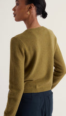 A woman shown from the back wearing Seasalt cropped, long-sleeve knit sweater in a muted olive green colour, paired with dark navy trousers, against a plain light background.