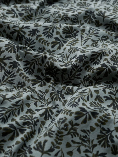 A close-up of draped fabric with an all-over botanical leaf pattern in dark olive green and black on a light gray background.