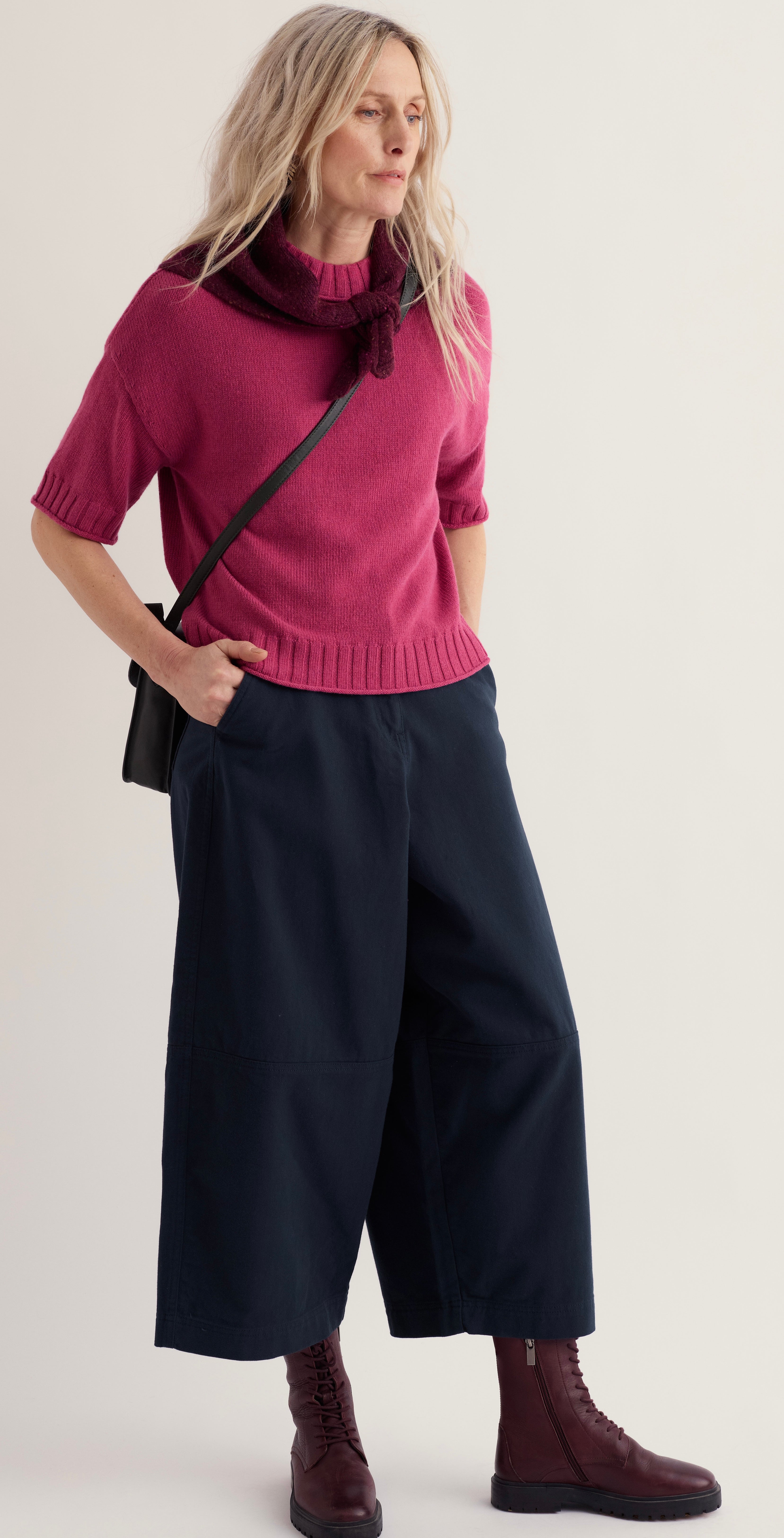 A woman modelling Seasalt short sleeve knit sweater in a dusty rose pink colour with a dark burgundy bow at the neck, paired with dark navy wide leg cropped pants and dark brown lace up boots, against a plain light grey background.
