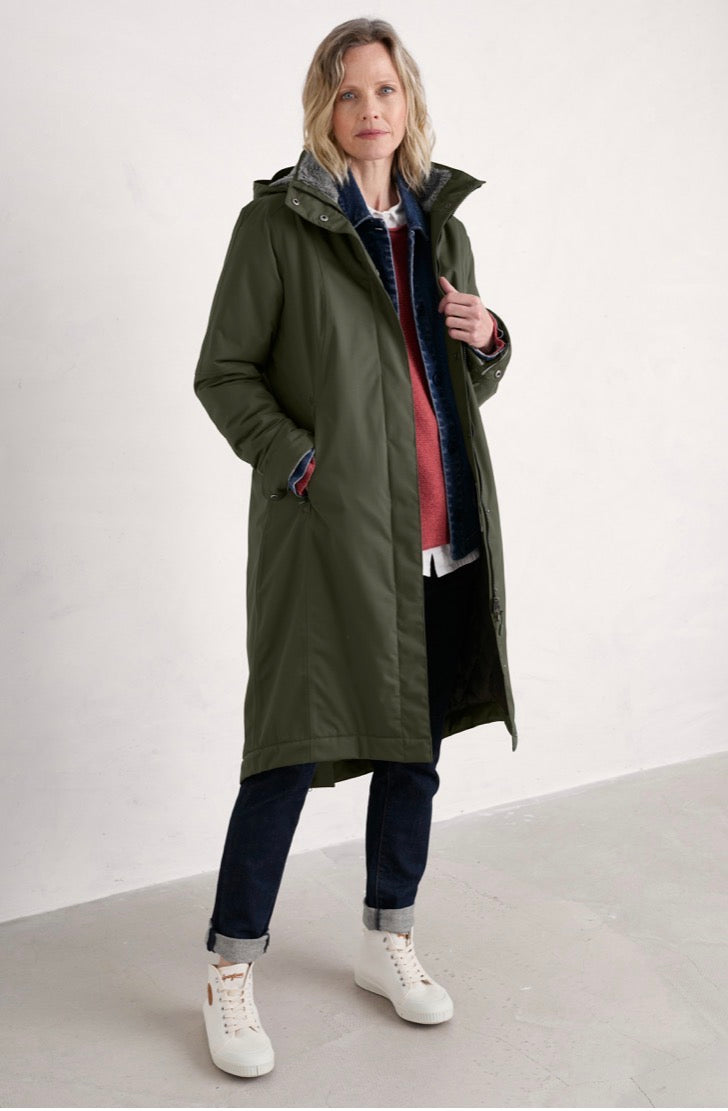 A woman modelling a Seasalt mid-length olive green hooded coat over a layered outfit, including a blue patterned shirt, red sweater, dark pants, and white sneakers, in a minimalist studio setting.
