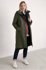 A woman modelling a Seasalt mid-length olive green hooded coat over a layered outfit, including a blue patterned shirt, red sweater, dark pants, and white sneakers, in a minimalist studio setting.