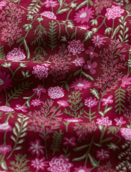 A close-up of a draped fabric with a dense floral and botanical print on a deep burgundy background, featuring pink flowers and muted sage green leaves.