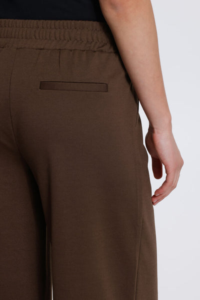Close up of a person wearing brown wide leg trousers, showing the back waistband, a side welt pocket, and the side of the leg against a light grey studio background.