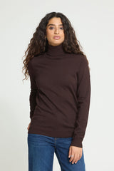 A woman with long, curly dark brown hair wearing a dark brown Ichi Rollneck sweater and blue jeans, standing against a plain light gray studio background.
