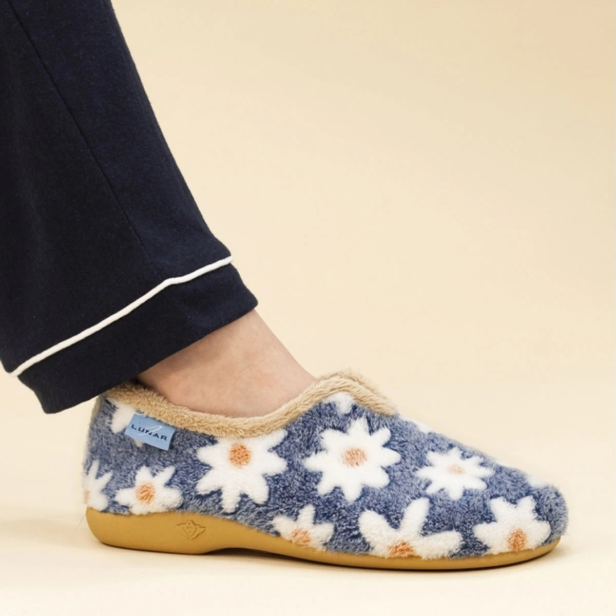 A person wearing a blue fuzzy slip-on slipper with a white daisy floral pattern and a tan sole, paired with navy pajama pants, against a light beige background.